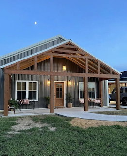 Rustic house exterior with covered porch, wooden beams, chairs, and grassy yard at dusk