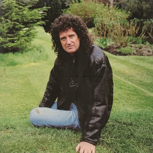 Man with curly dark hair in black leather jacket crouching on green lawn outdoors