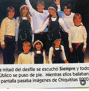 Children posing on stage in white shirts and dark overalls, with Spanish text below