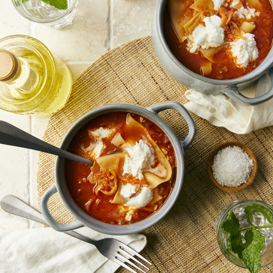 Overhead view of two bowls of lasagna soup garnished with dollops of ricotta cheese, accompanied by olive oil and herbs
