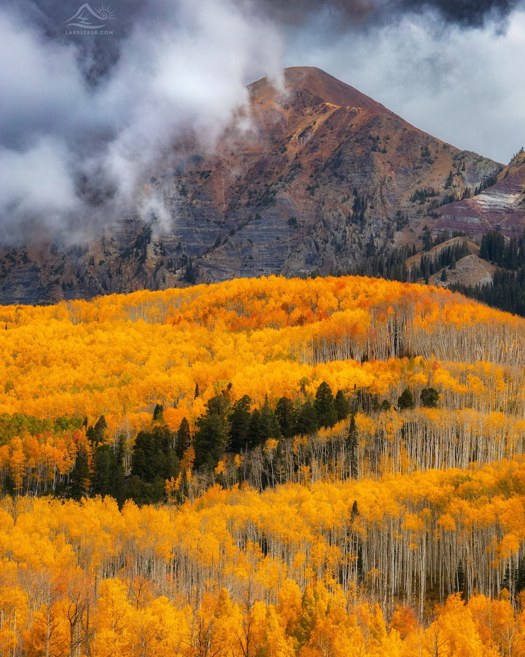 Golden autumn aspens on rolling hills below a rocky mountain peak and clouds