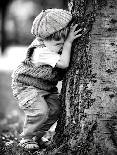 Black and white photo of a small child in knit hat and sweater hiding face against tree trunk