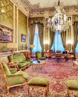 Opulent palace room with green velvet chairs, crystal chandelier, gold trim walls, and Persian carpet