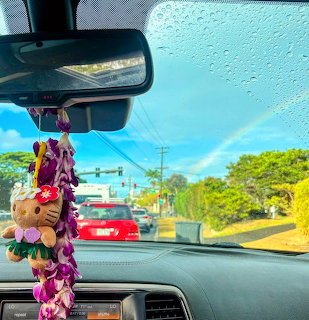 Rainbow over Hawaiian road seen through car windshield with colorful lei hanging from mirror