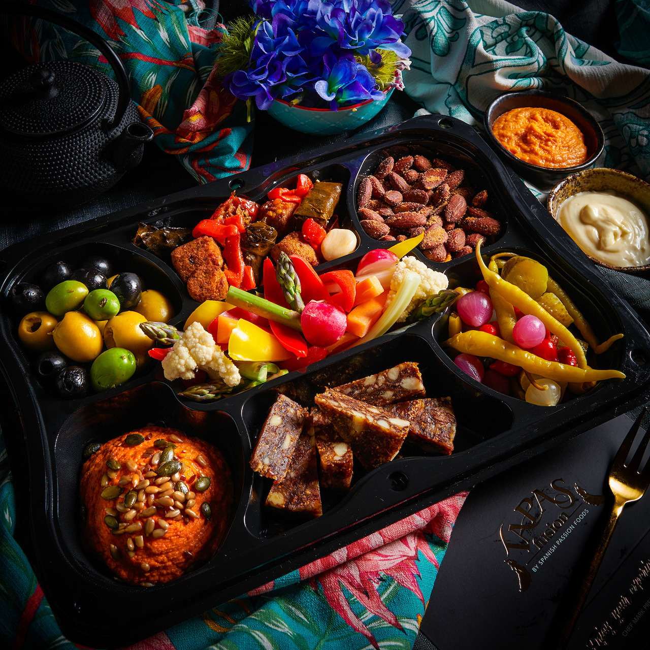 Vegan tapas platter with olives, vegetables, bread, dips, and pickled peppers on a dark table