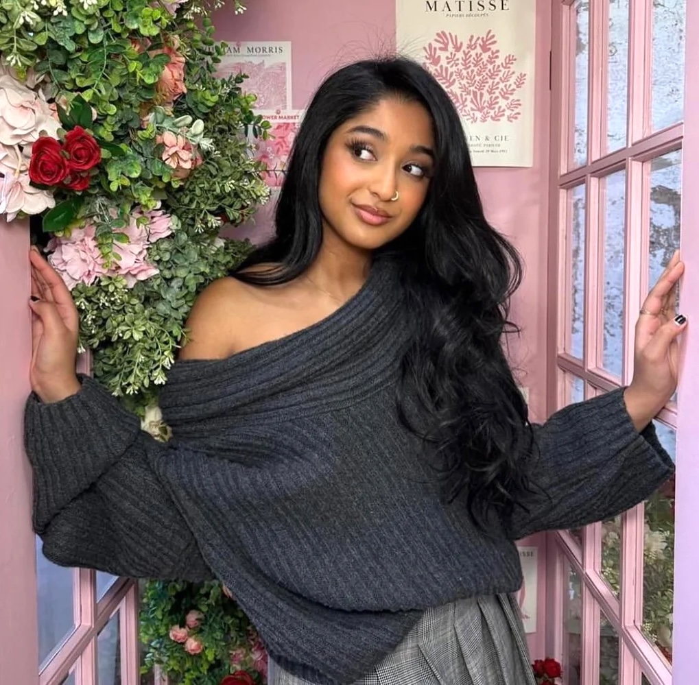 Maitreyi Ramakrishnan posing in a pink doorway beside flowers in a gray off-shoulder sweater