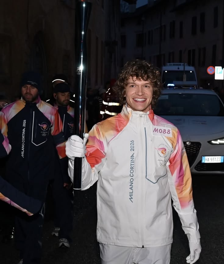 Smiling young man holding an Olympic torch at night in Milano Cortina 2026 uniform
