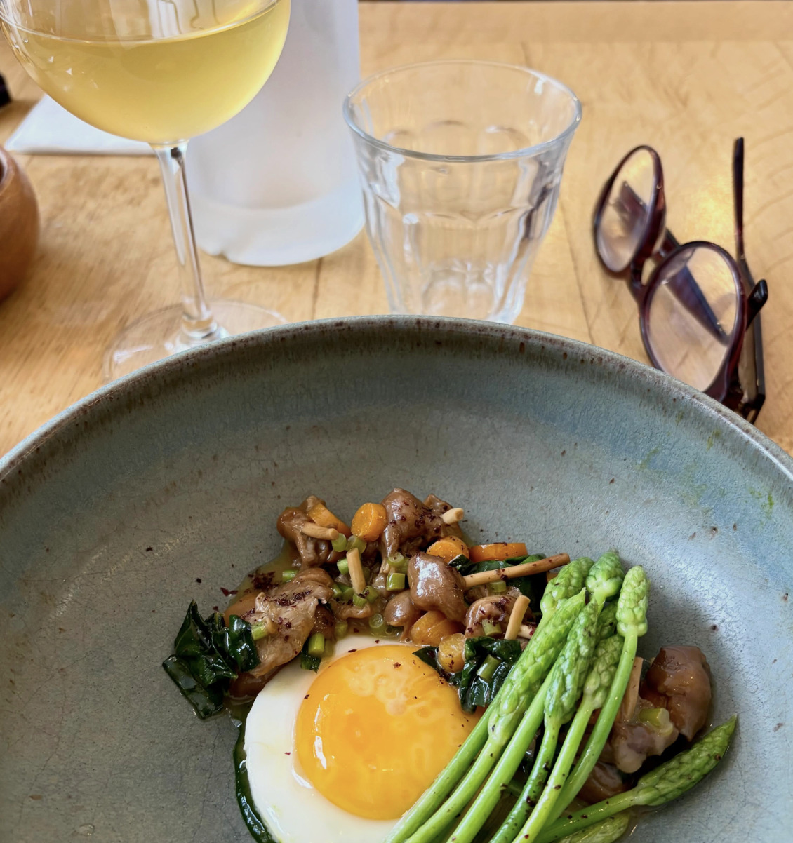 Ceramic bowl with fried egg, asparagus, sautéed mushrooms and greens, with white wine glass on wooden table