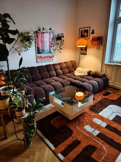 Retro 70s living room with brown sectional sofa, orange shag rug, plants, and vintage lighting