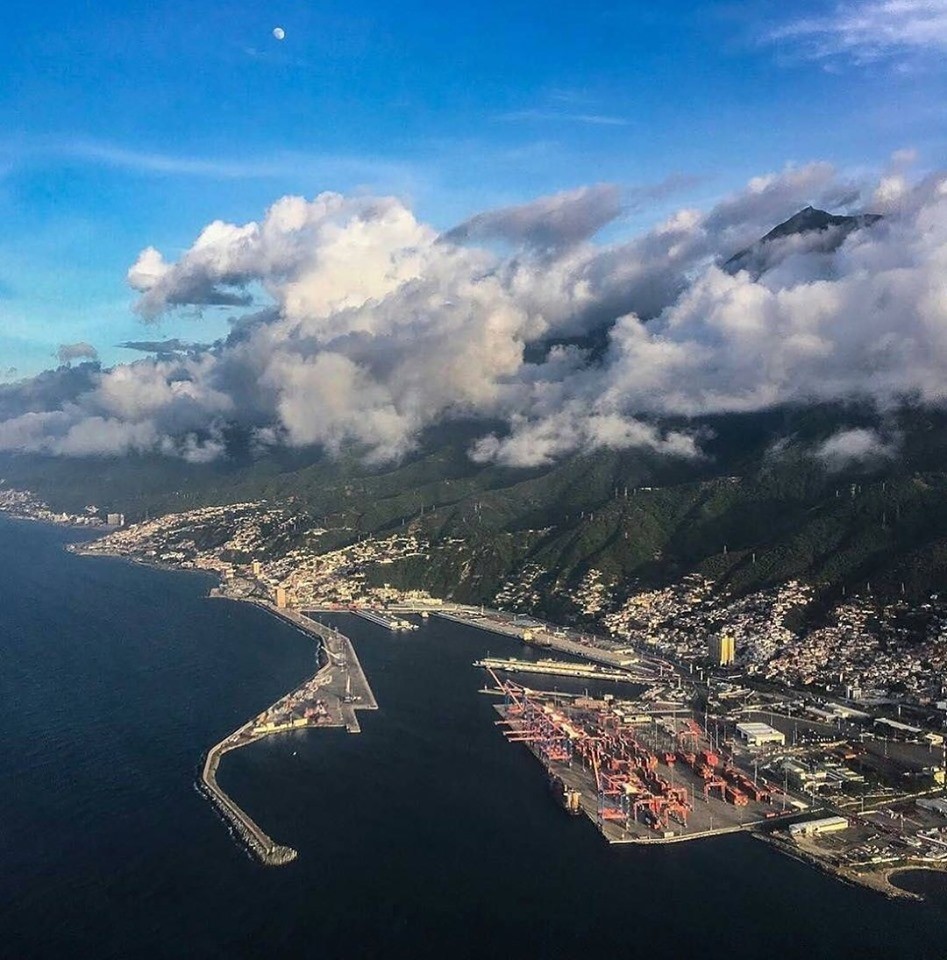Aerial view of La Guaira harbor, coastal city, and green mountains under large clouds