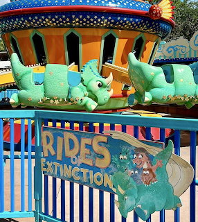 Colorful dinosaur-themed ride cars behind a blue fence at a theme park