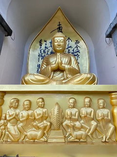 Golden Buddha statue with praying hands above a carved panel of smaller figures in a temple interior