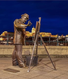 Bronze statue of a painter working at an easel on a promenade at night with lit buildings behind