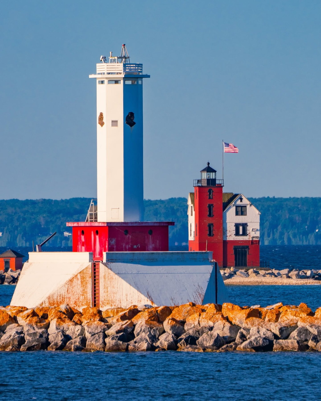 White lighthouse with red buildings on a rocky breakwater beside the water under a blue sky