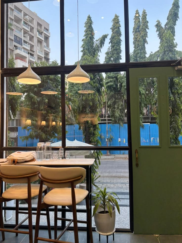 Cafe interior view through window showing tables, pendant lights, and outdoor garden with blue planters