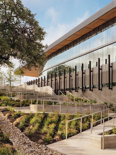 Modern arena building with curved wood roof, glass walls, outdoor stairs and landscaping