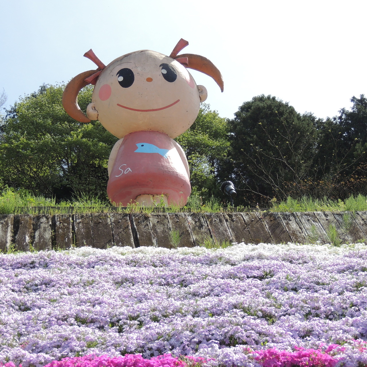 Smiling cartoon mascot character behind colorful carpet of pink and purple moss phlox flowers
