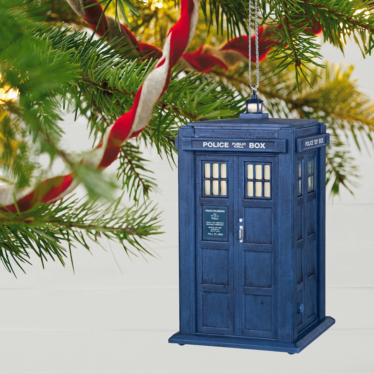 Blue TARDIS police box ornament hanging on Christmas tree with lights and ribbon