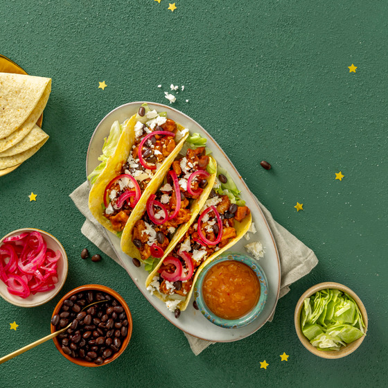 Three chicken tacos with pomegranate seeds, cheese, and cilantro on a white plate with side bowls of toppings