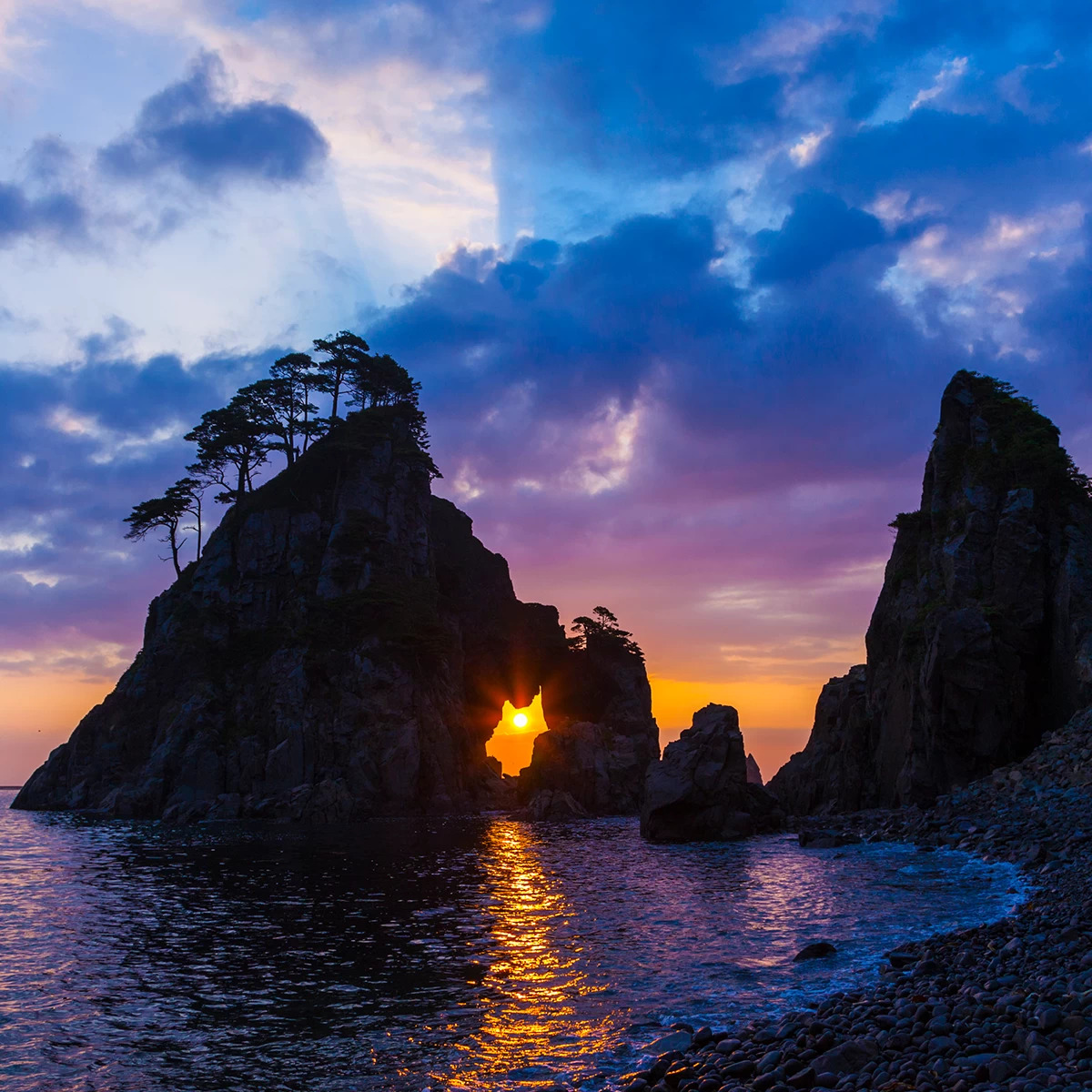 Sunset glowing through rocky sea stacks on a calm coastline under purple-blue clouds