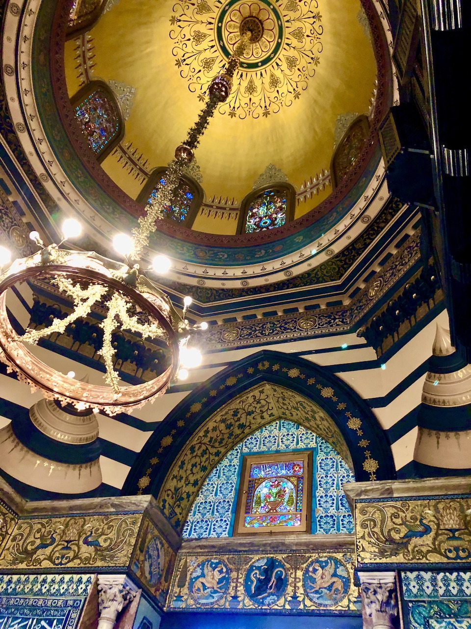 Ornate interior with a golden dome, chandeliers, blue arches, and mosaic tilework