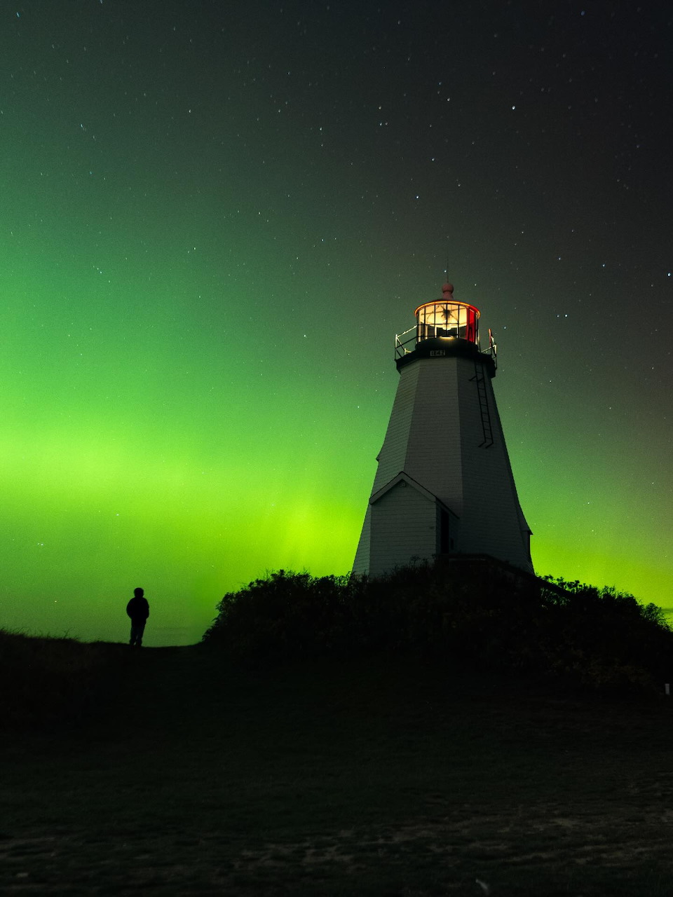 Silhouetted lighthouse with glowing light against green aurora borealis and starry night sky