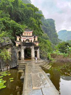 Vietnamese temple entrance with ornate gate, stone bridge over pond, and karst mountains behind under cloudy sky