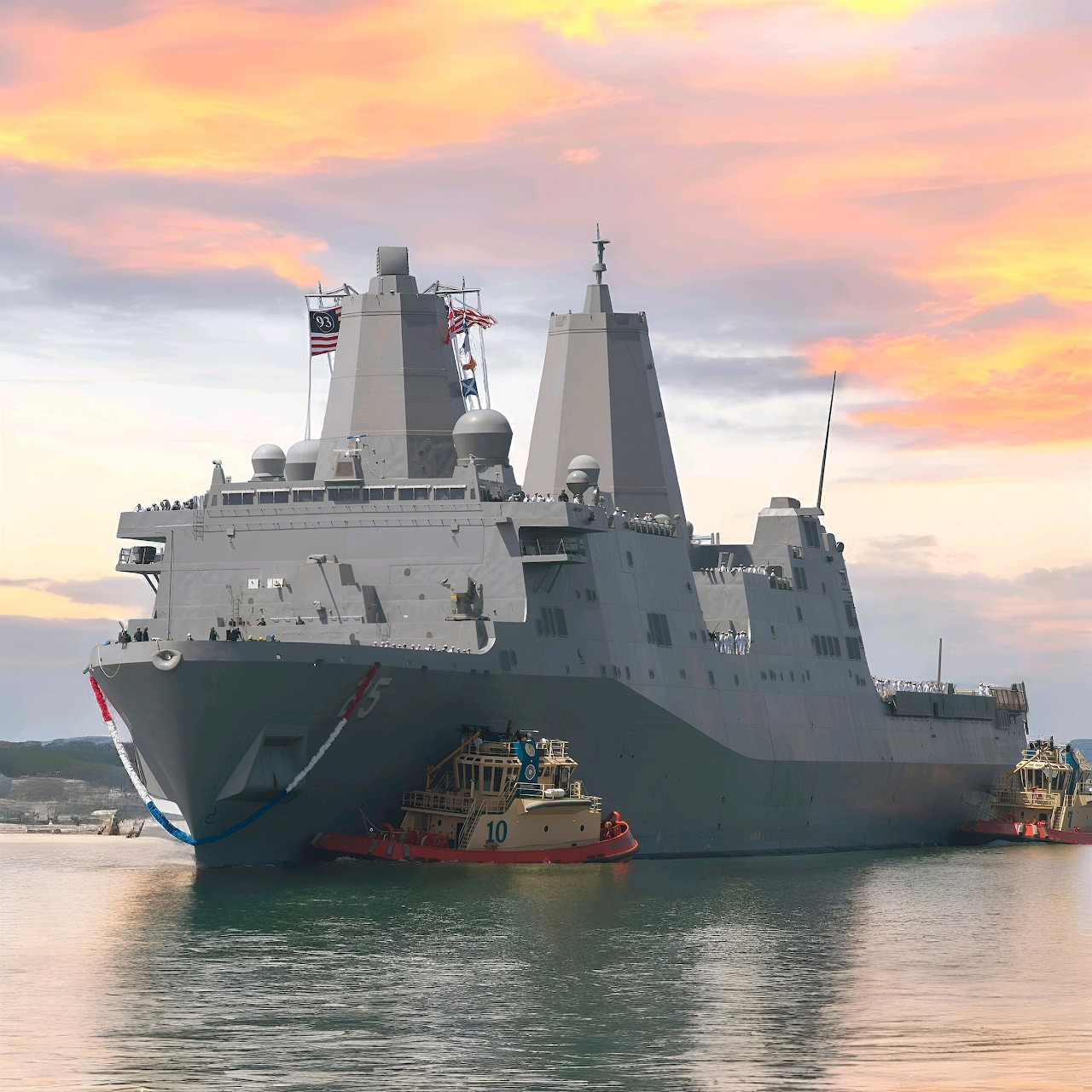 USS San Antonio naval ship docked on calm water at sunset with a small tugboat nearby