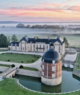 Aerial view of a French chateau with round tower, moat, and green grounds under a misty sunrise sky