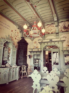 Ornate historic interior with carved chairs, chandeliers, painted walls, and decorative woodwork