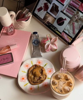 Pink aesthetic desk with iced coffee, cookie on floral plate, hand cream, tablet showing makeup, and pink accessories