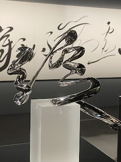 Shiny chrome steel sculpture in the form of Chinese calligraphy on a white pedestal in gallery