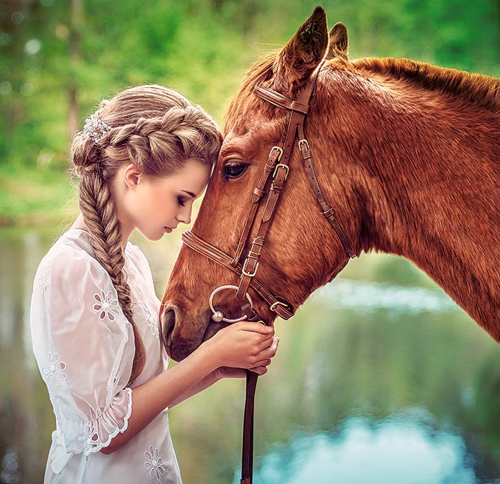 Young girl in a white dress touching a brown horse by a pond
