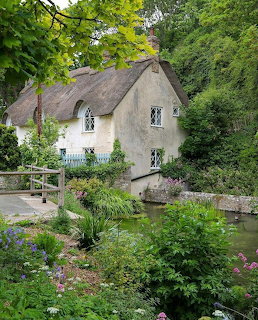 White thatched cottage beside a pond with lush garden greenery and flowers