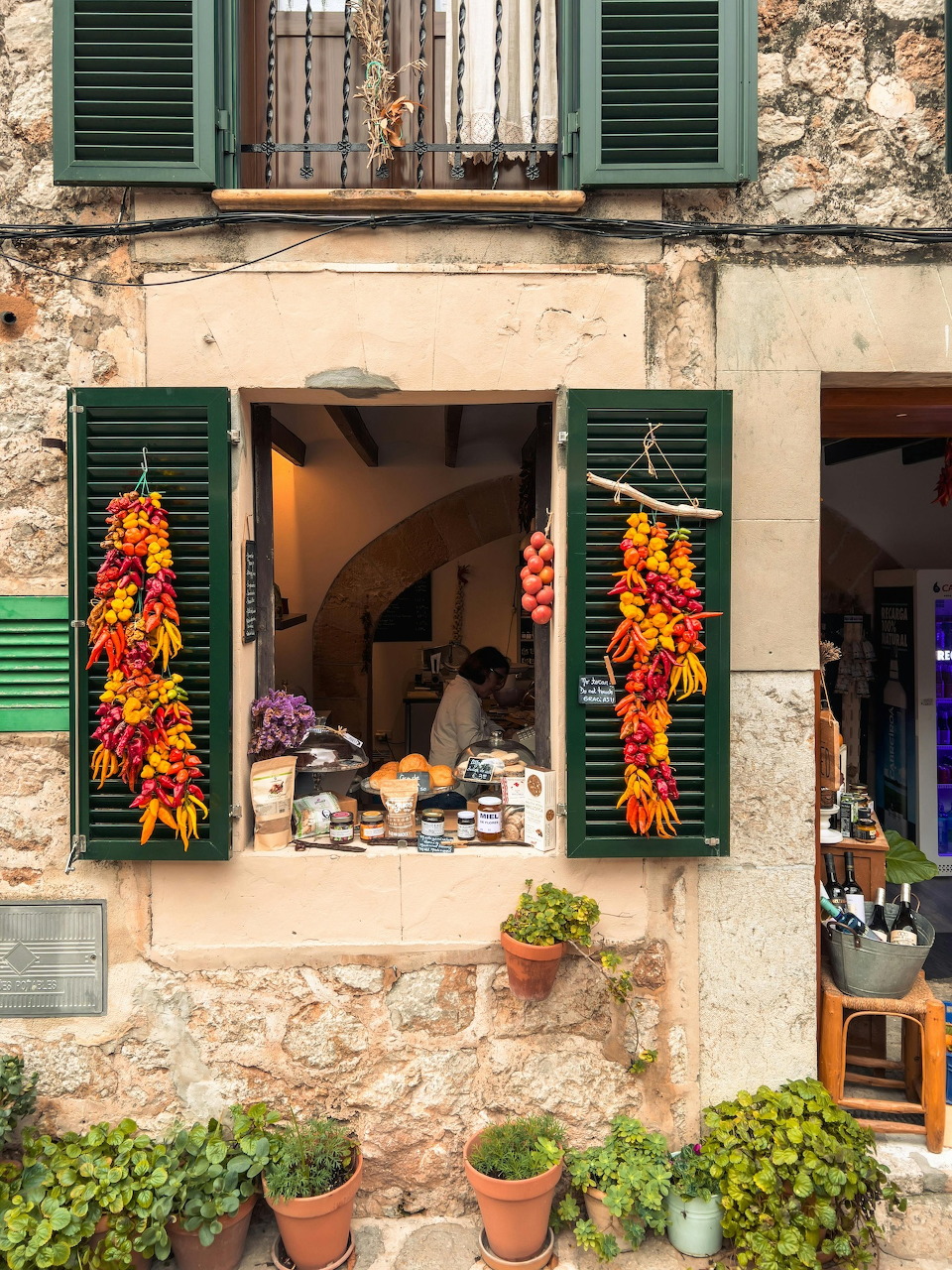 Rustic stone shop facade with green shutters, potted plants, and strings of red and yellow peppers