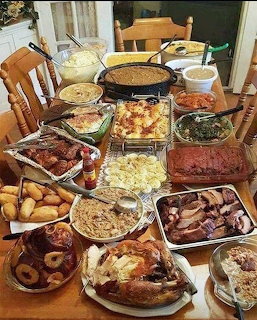 Indoor dining table filled with buffet dishes, roasted meats, potatoes, pasta, greens, and casseroles
