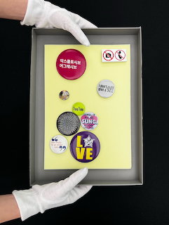 White-gloved hands holding a framed set of colorful pin badges on a yellow background