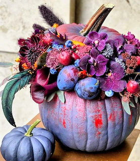 Blue-painted pumpkin with purple floral arrangement including orchids, plums, and dark berries on a wooden table