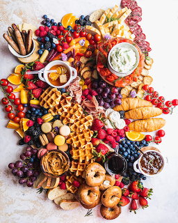Colorful breakfast charcuterie board with waffles, bagels, berries, grapes, sausage and spreads on marble surface