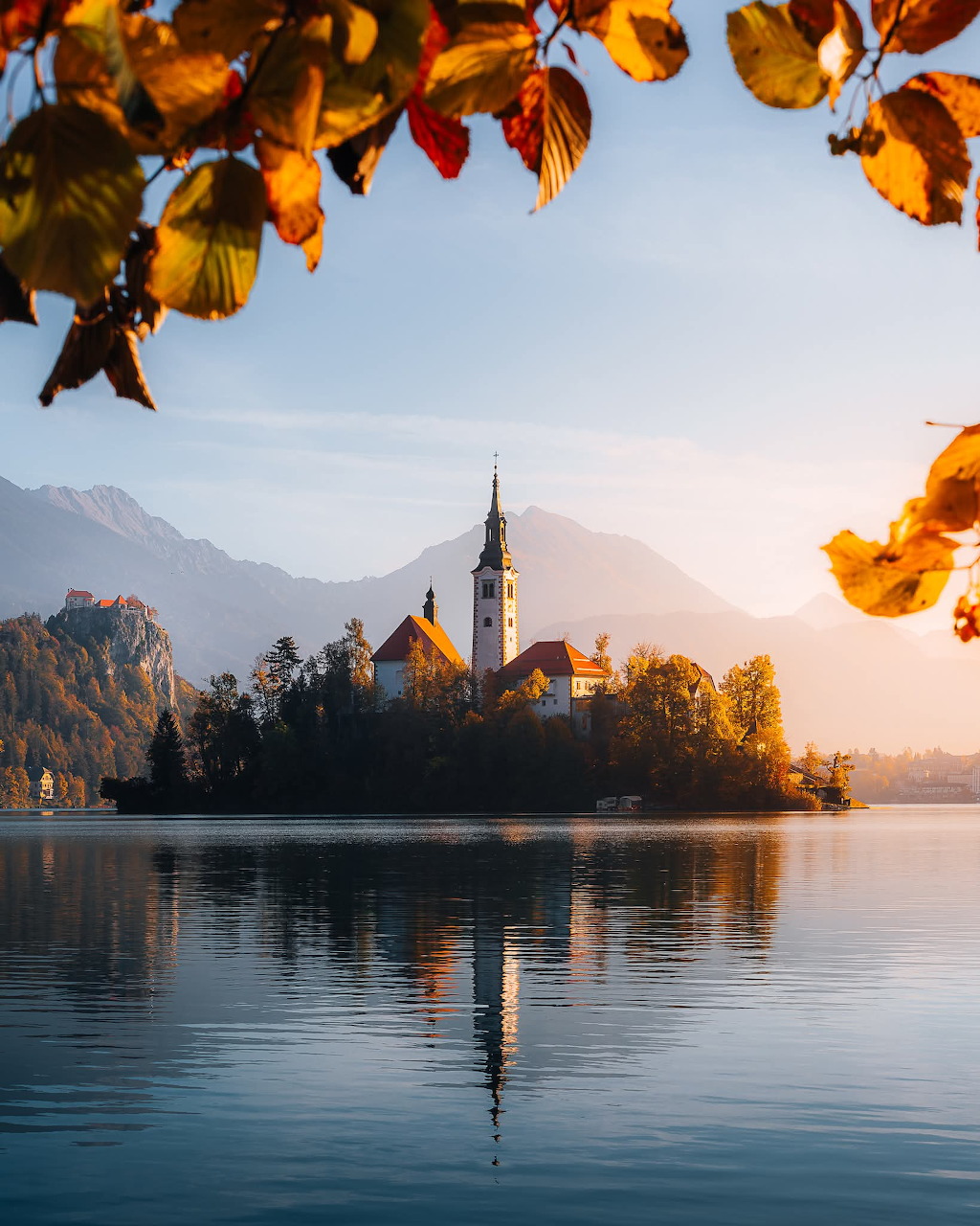 Lake Bled island church reflected in calm water, framed by autumn leaves and mountain backdrop, Slovenia