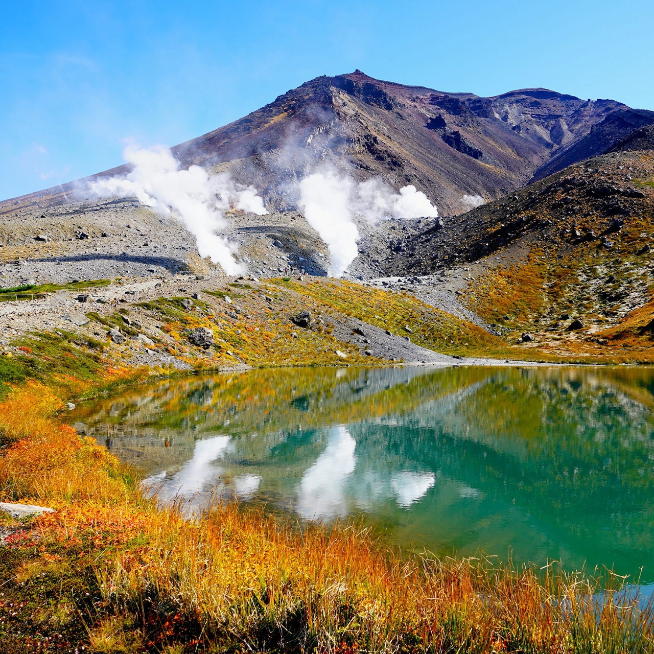 Rocky mountain with steam vents reflected in a calm green lake, bordered by autumn grasses
