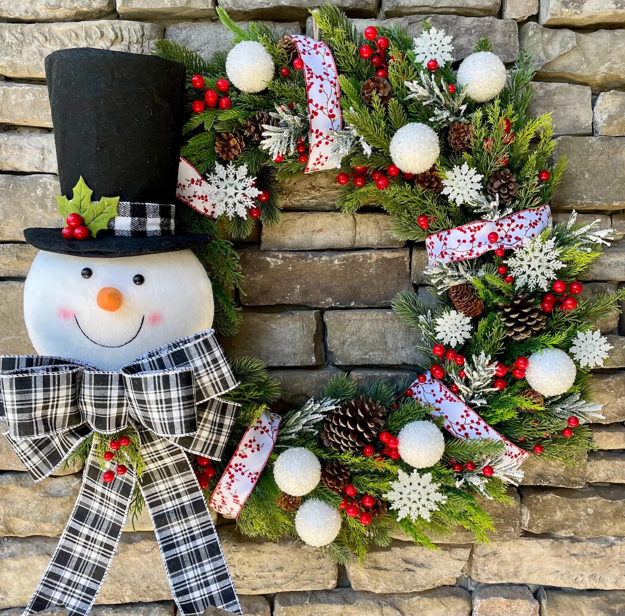 Christmas wreath with snowman decoration, white balls, red berries, pine greenery, and checkered scarf on stone background
