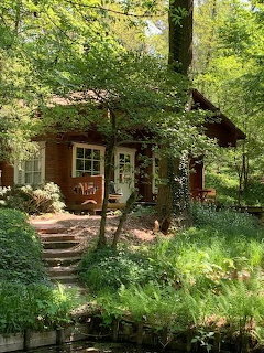 Red wooden cabin surrounded by dense green trees and garden plants with a stone path