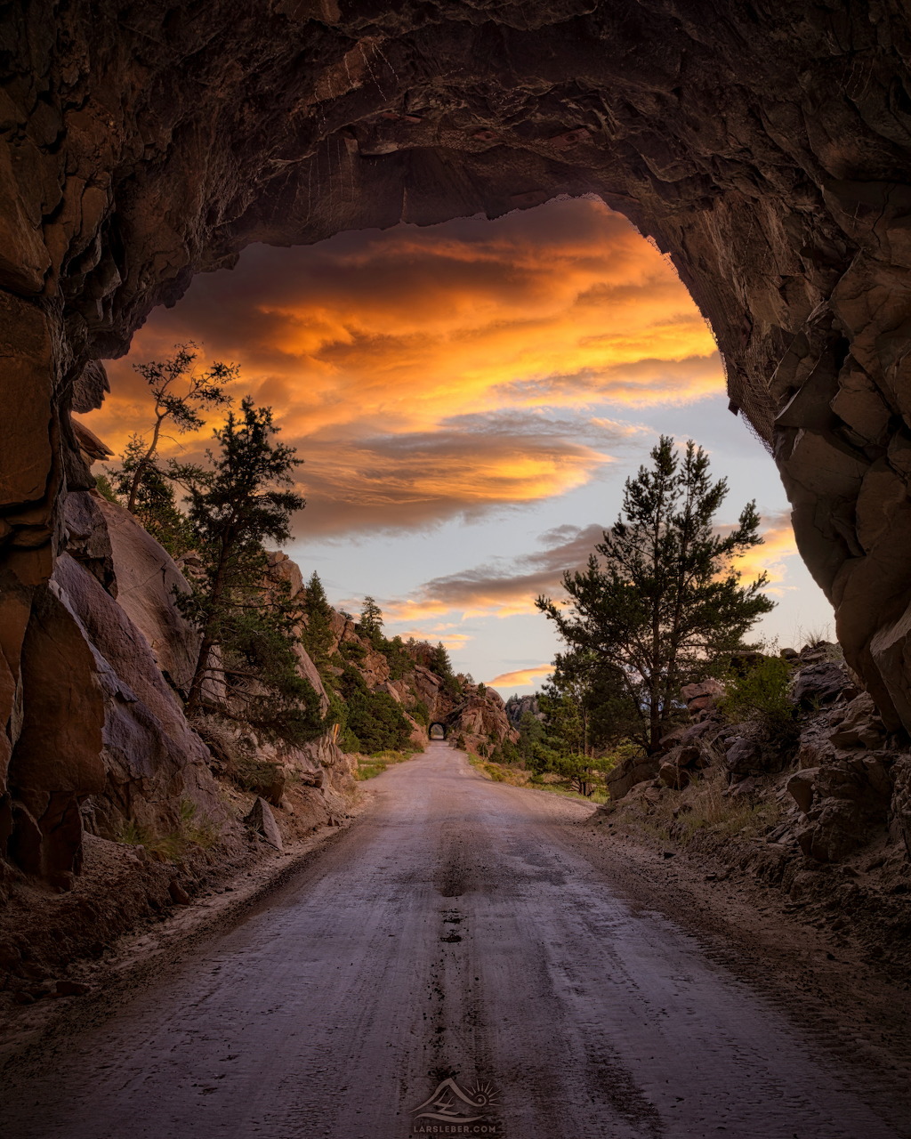 View through a rocky tunnel arch onto a dirt road with a vivid orange sunset sky and silhouetted pine trees.