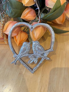 Silver heart ornament with two birds on a wooden surface beside orange flowers