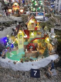 Lit miniature yellow house with red roof in snowy Christmas village scene with fountain and trees