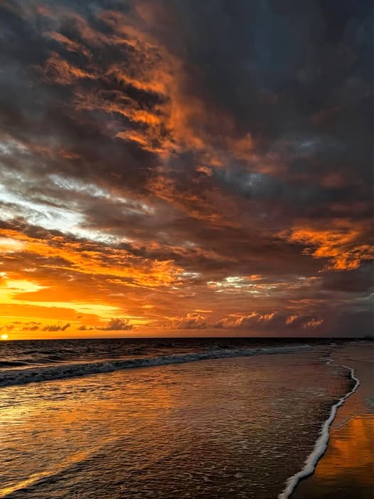 Stormy sunset over a beach with orange clouds, wet sand reflections, and a gentle wave at the shore