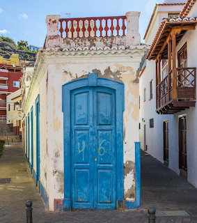 Weathered white building with bright blue wooden door and ornate balcony railing in narrow village street