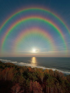 Three concentric rainbows spanning across sky above ocean at sunset with sun on horizon