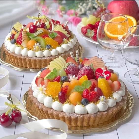 Two fruit tarts decorated with colorful fresh fruits and whipped cream on a table with glasses and cheries